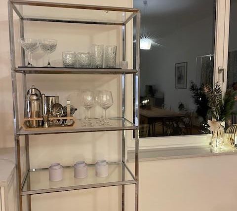 Ein stilvoller Glasregal mit Gläsern und einem Kännchen steht vor einem Fenster. Der Raum ist warm beleuchtet und hat eine gemütliche Atmosphäre.