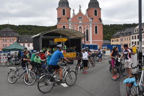 Eine Gruppe von Radfahrern versammelt sich vor einer historischen Kirche. Im Hintergrund sind Gebäude und eine Bühne zu sehen.