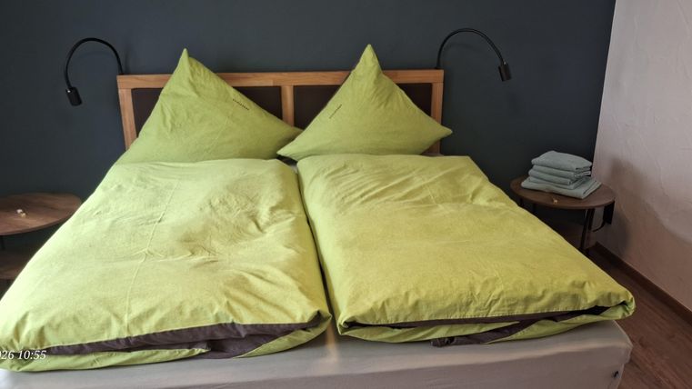 A cozy bedroom with a double bed and two green pillows. Next to the bed is a small table with a lamp and fresh towels.