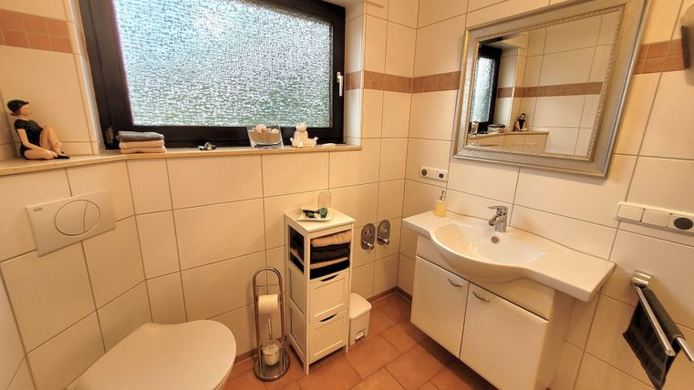 Ein modernes Badezimmer mit einer hellen Fliese und einem großen Spiegel. Es gibt eine Toilette, ein Waschbecken und ein Fenster, das Tageslicht hereinlässt.