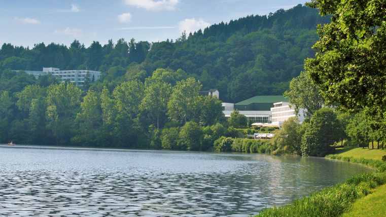 Seeufer des Stausees Bitburg mit bewaldeten Hügeln und Gebäuden im Hintergrund.