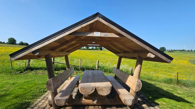 Un abri en bois avec une grande table et des bancs se trouve sur une prairie. En arrière-plan, on peut voir des champs verts et un ciel bleu clair.