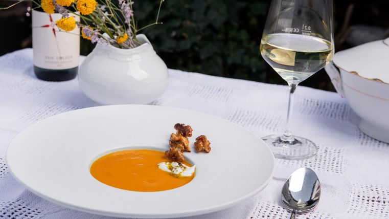 Een elegante soep in een wit bord, gegarneerd met Eifelfrisse en een dot room. Ernaast staat een glas witte wijn en een kleine vaas met bloemen.