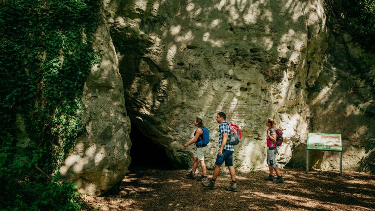 Drie wandelaars betreden een grot in een bebost gebied. De omgeving is groen en omringd door bomen.