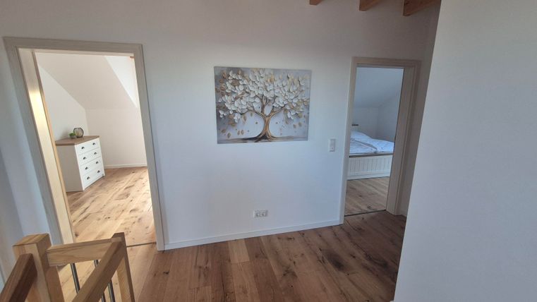 Un couloir lumineux avec un sol en bois et des poutres en bois au plafond. Deux portes mènent à des chambres adjacentes, et au mur se trouve un tableau d'un arbre.