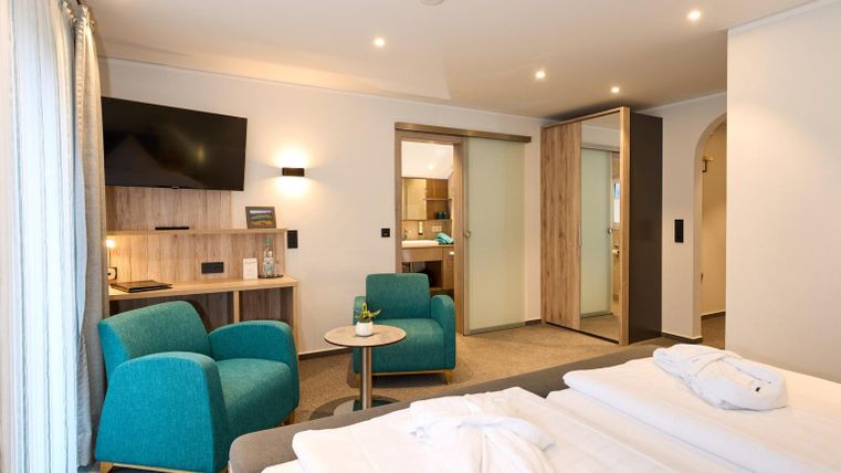 Een moderne hotelkamer met twee eenpersoonsbedden. Er is een kleine zithoek met blauwe stoelen en een tafel, evenals een televisie aan de muur.
