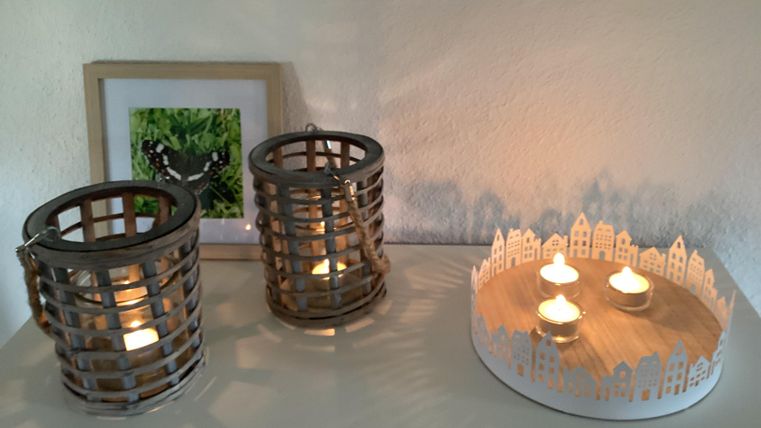 Zwei dekorative Laternen mit Kerzen stehen auf einem Tisch. Daneben befindet sich ein Bilderrahmen und eine runde Kerzenhalterung mit kleinen Lichthäusern.