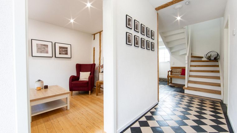 Ein heller Flur mit einem schachbrettartigen Bodenbelag und einer Treppe. Auf der linken Seite befindet sich ein roter Sessel und einige Bilder an der Wand.