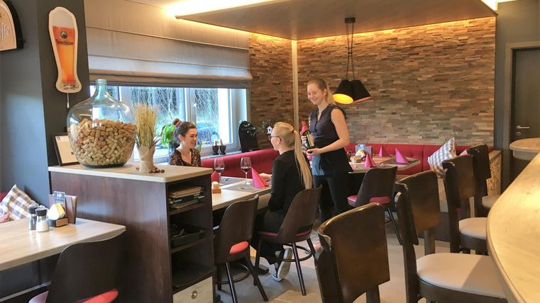 Een modern restaurant met een gezellige sfeer. Gasten zitten aan tafels, terwijl een ober hen vriendelijk begroet.