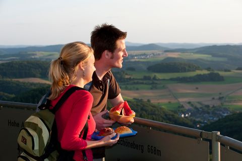Ein Paar steht auf einem Aussichtsturm und blickt in die Ferne. Sie halten Snacks in den Händen. Im Hintergrund ist eine hügelige Landschaft zu sehen.