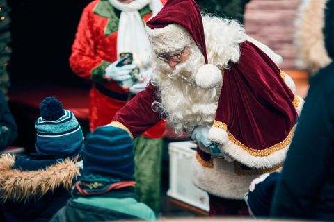 Ein Weihnachtsmann verteilt Geschenke an Kinder. Im Hintergrund sind festlich gekleidete Personen zu sehen.