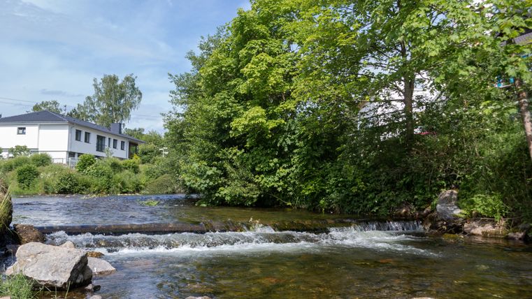 Une petite rivière à l'eau peu profonde et aux pierres, entourée d'arbres verts et d'une maison blanche en arrière-plan.