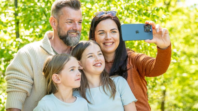Une famille heureuse fait un selfie en plein air. En arrière-plan, on peut voir des arbres verts et beaucoup de lumière du soleil.