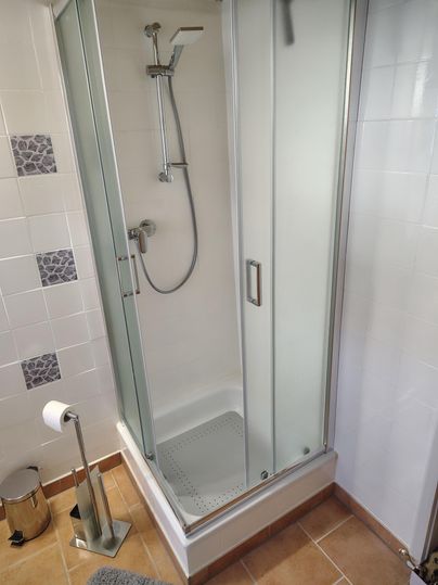 A modern shower with glass front and handheld showerhead. The floor is tiled and there is a towel holder as well as a toilet paper roll.
