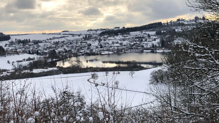 Eine verschneite Landschaft mit einem ruhigen See und einem kleinen Dorf im Hintergrund. Der Himmel ist bewölkt und die Umgebung wirkt friedlich und winterlich.