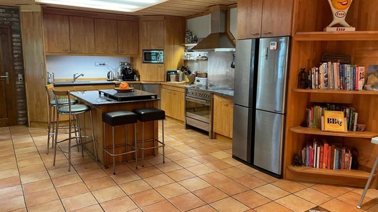 A modern kitchen with wooden furniture and a large window. The room is well-lit and has a dining area with tall chairs.