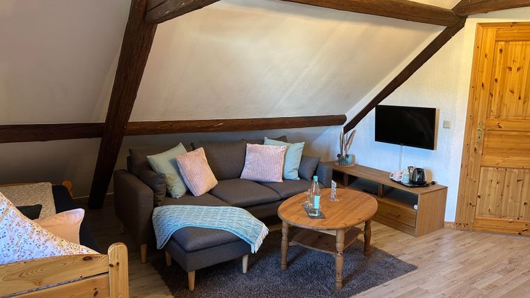 Een gezellige woonruimte met een grijze bank, kleurrijke kussens en een ronde tafel. Aan de muur hangt een televisie, en er is een houten deur.