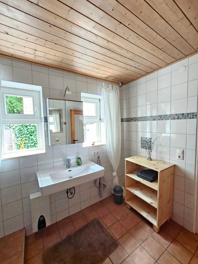 Een lichte badkamer met witte tegels en een houten plafond. Er is een wastafelgebied en een houten rek met handdoeken.