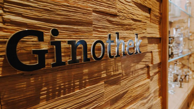 Een houten wand met de tekst "Ginothek" in een modern ontwerp. Op de achtergrond zijn glazen planken met verschillende gin-flessen zichtbaar.