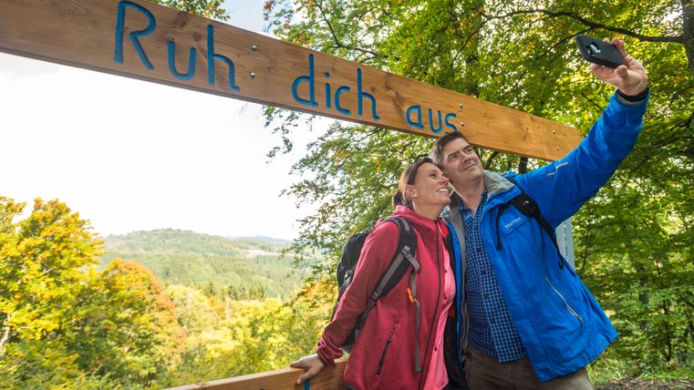 Un couple prend un selfie sous un panneau en bois portant l'inscription 'Repose-toi' dans un paysage forestier verdoyant.