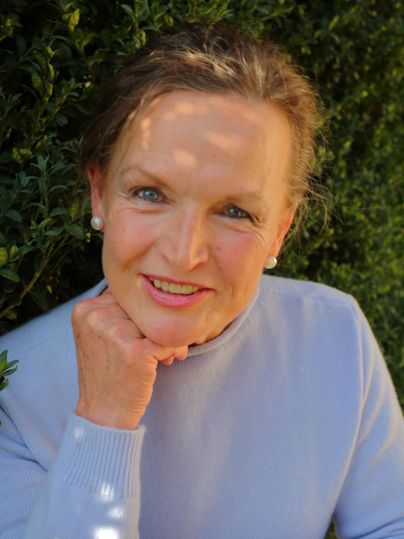 Eine Frau mit lächelndem Gesicht sitzt vor einer grünen Hecke. Sie trägt einen hellblauen Pullover und hat eine Hand an ihrem Kinn.
