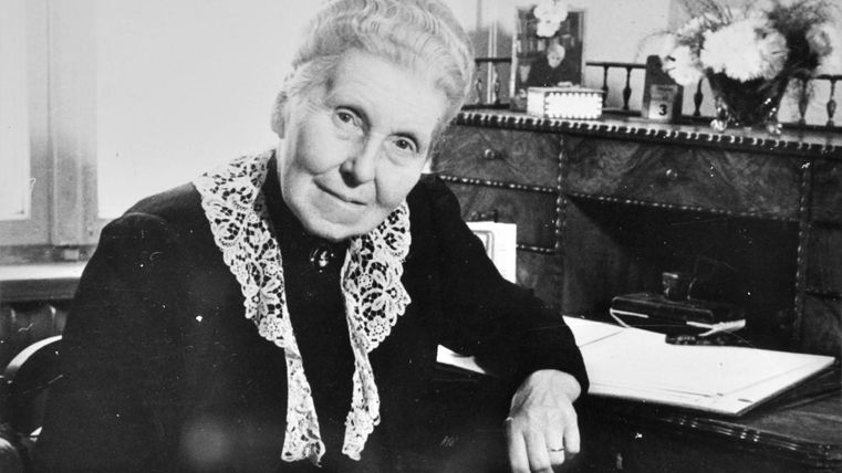 Eine ältere Frau sitzt an einem Schreibtisch und lächelt in die Kamera. Im Hintergrund ist eine Vase mit Blumen und einige persönliche Gegenstände zu sehen.