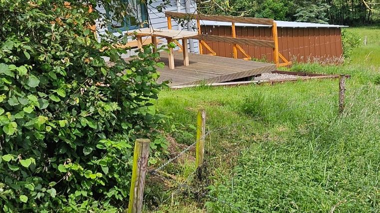 A wooden terrace with a railing, surrounded by greenery and trees. In the background, a building is visible.