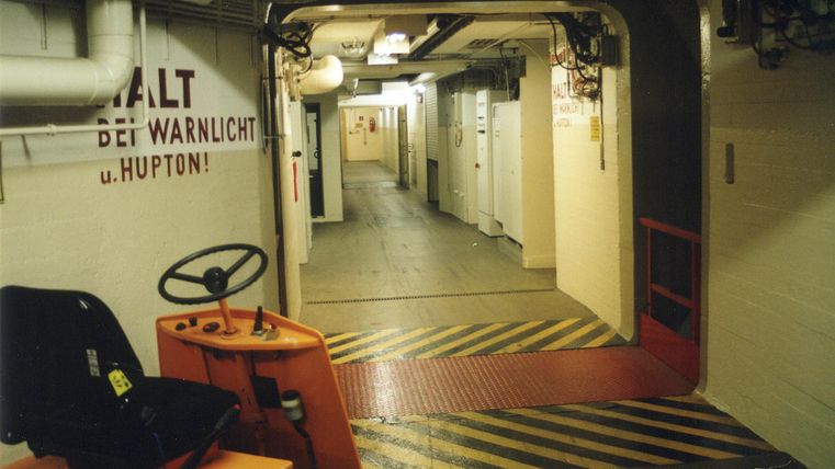 Ein langer Gang in einem modernen Gebäude mit einem roten Boden und gelben Streifen. Auf der linken Seite steht ein Wagen mit einem Lenkrad.
