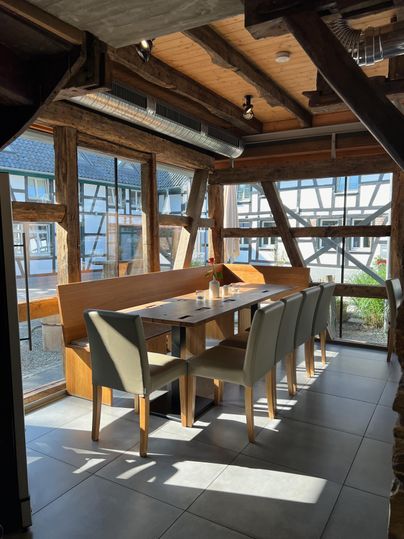 Ein modernes Restaurant mit einem großen Tisch und eleganten Stühlen. Große Fenster bieten viel natürlichem Licht und einen Blick nach draußen.