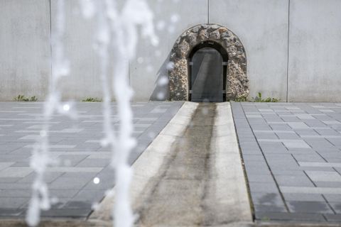 Ein moderner Platz mit grauen Fliesen und einem interessanten Wasserlauf. Im Hintergrund sieht man einen steinernen Bogen.
