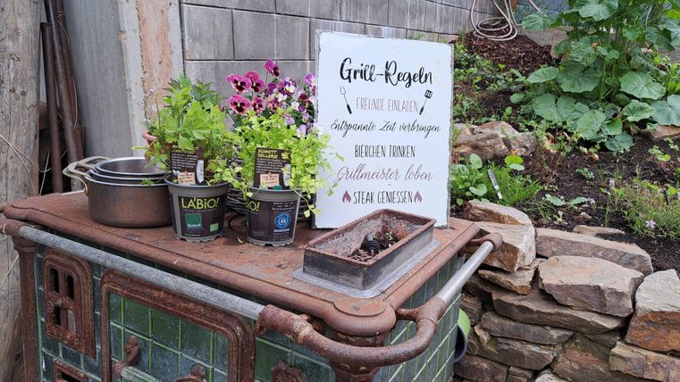 Ein alter Ofen mit Blumenkästen und einer Tafel, die Anweisungen für das Gießen von Pflanzen gibt. Im Hintergrund sind grüne Pflanzen sichtbar.