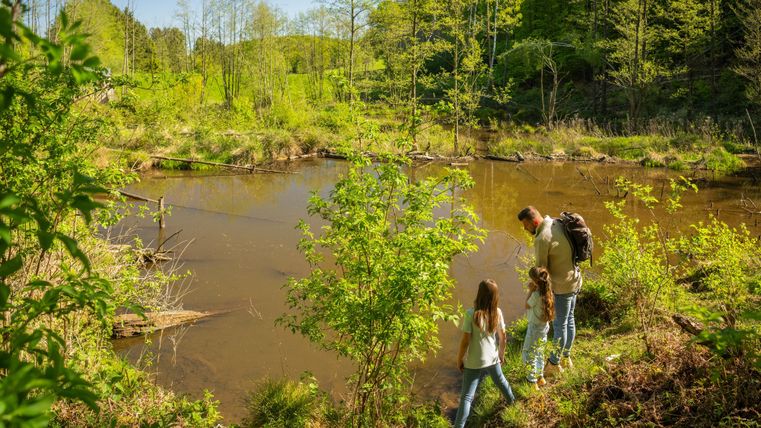 Une famille se tient près d'un plan d'eau calme, entourée d'une végétation luxuriante. Le soleil brille et c'est une belle journée en pleine nature.