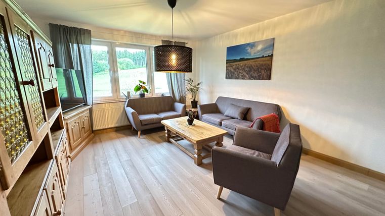 Ein modernes Wohnzimmer mit grauen Sofas und einem Holztisch. Große Fenster bringen viel Licht herein und bieten einen Blick ins Freie.