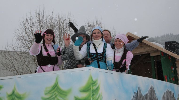 Eine Gruppe von Menschen in festlicher Kleidung steht auf einem Wagen. Es schneit leicht und die Atmosphäre ist fröhlich.