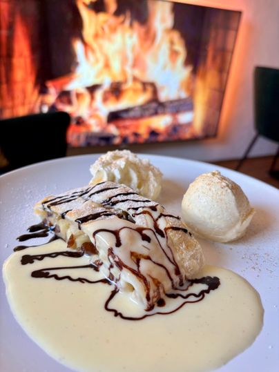 A delicious dessert with apple strudel, cream, and ice cream is on a plate. In the background, a cozy fire is burning.