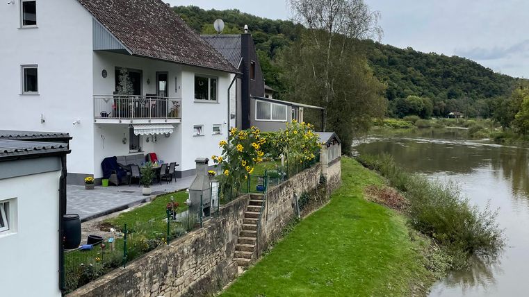 Ein schönes Haus am Fluss mit grünem Rasen und Bäumen im Hintergrund. Der Himmel ist bewölkt und verleiht der Szene eine ruhige Atmosphäre.
