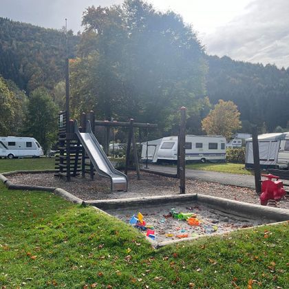 Un terrain de jeux avec un toboggan et une sable. En arrière-plan, on peut voir des caravanes et des arbres.