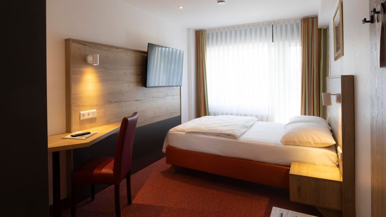 A modern hotel room with a double bed and a desk. Large windows provide plenty of daylight.