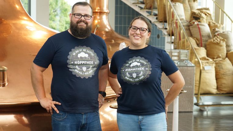 Twee personen staan glimlachend voor een koperen brouwapparaat. Beide dragen T-shirts met de opdruk "HOPPENHEK".