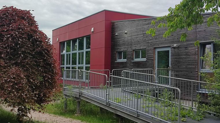 Un bâtiment moderne avec une façade rouge et lambrissée en bois. Au premier plan, on aperçoit une rampe pour un accès sans barrières.