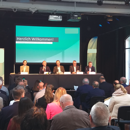 Een conferentie met meerdere sprekers aan een tafel voor een publiek. Op de achtergrond is een scherm te zien met de tekst "Hartelijk welkom!"