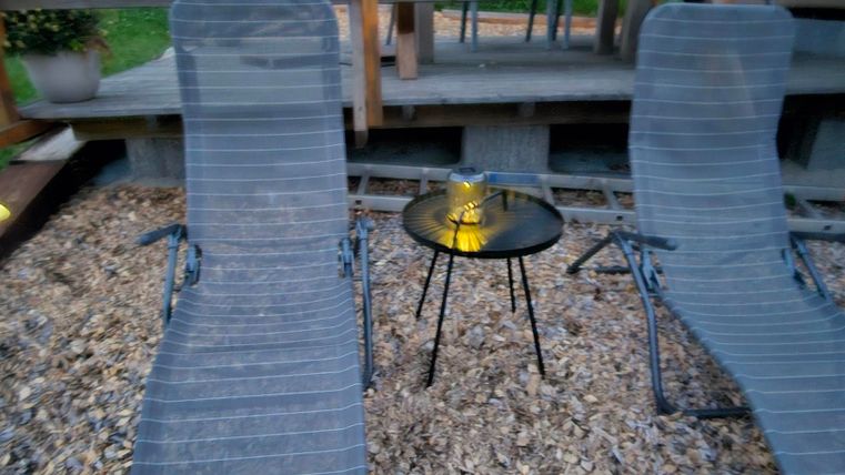 Two lounge chairs are located on a gravel floor next to a small table. In the background, a wooden terrace can be seen.