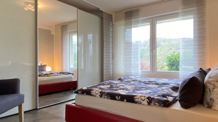 A cozy bedroom with a large bed and a mirror. The windows offer a view outside and let in a lot of daylight.