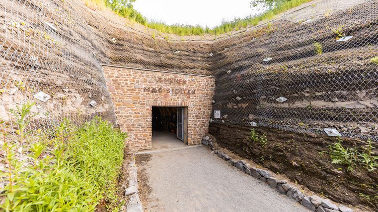 Ein Eingang aus Stein führt in einen Tunnel. Der Weg dorthin ist mit Kies bedeckt. Links und rechts stehen hohe Steinmauern. Oben wächst grünes Gras.