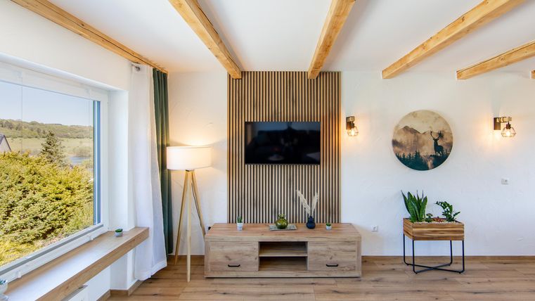 A modern living room with wooden ceiling beams and a television on the wall. There are light furniture and plants that create a cozy atmosphere.