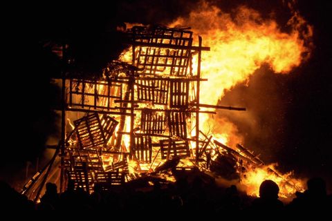 A large fire is burning in a destroyed wooden structure. People are standing nearby and watching the inferno.