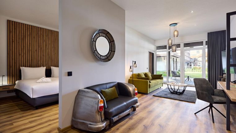 Une chambre moderne avec un coin salon confortable et des meubles élégants. De grandes fenêtres offrent une vue sur un jardin verdoyant.