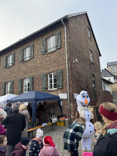 Eine festliche Veranstaltung vor einem alten Gebäude mit einem Kostüm von einem Schneemann. Viele Menschen, darunter Kinder, stehen zusammen und genießen die Atmosphäre.