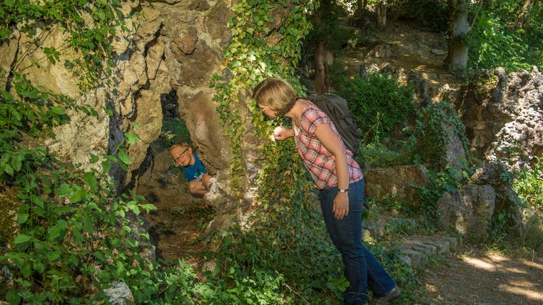 Een vrouw met een rugzak kijkt in een met klimop begroeide rotsopening waarin een kind zit.
