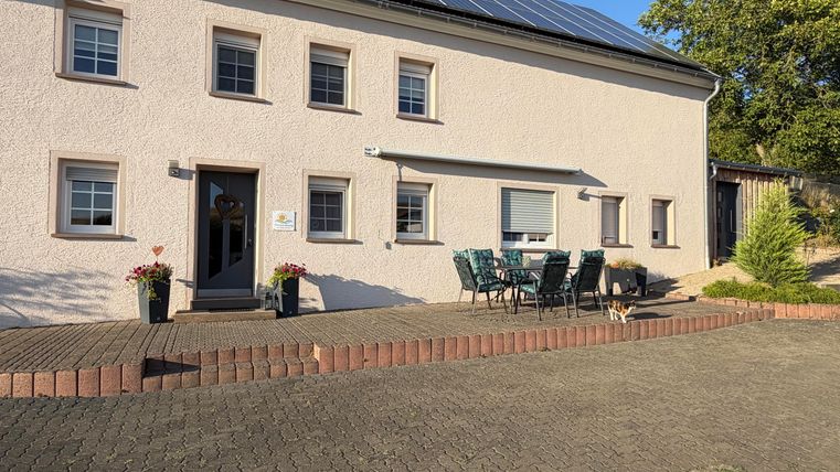 Een modern, wit huis met zonnepanelen op het dak. Voor het huis bevindt zich een terras met tuinstoelen en bloemen.